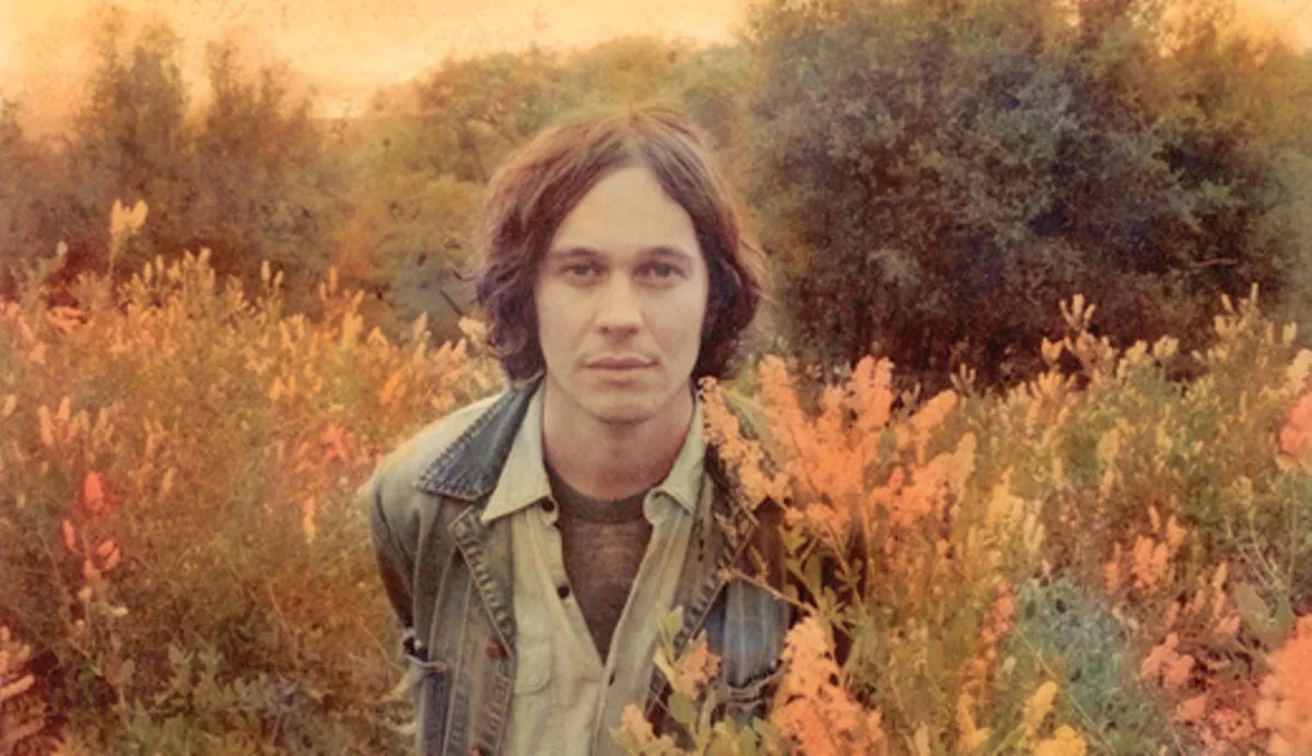 Ernest Greene, a man with long dark hair in a denim jacket in a field of yellow, red, and green plants.
